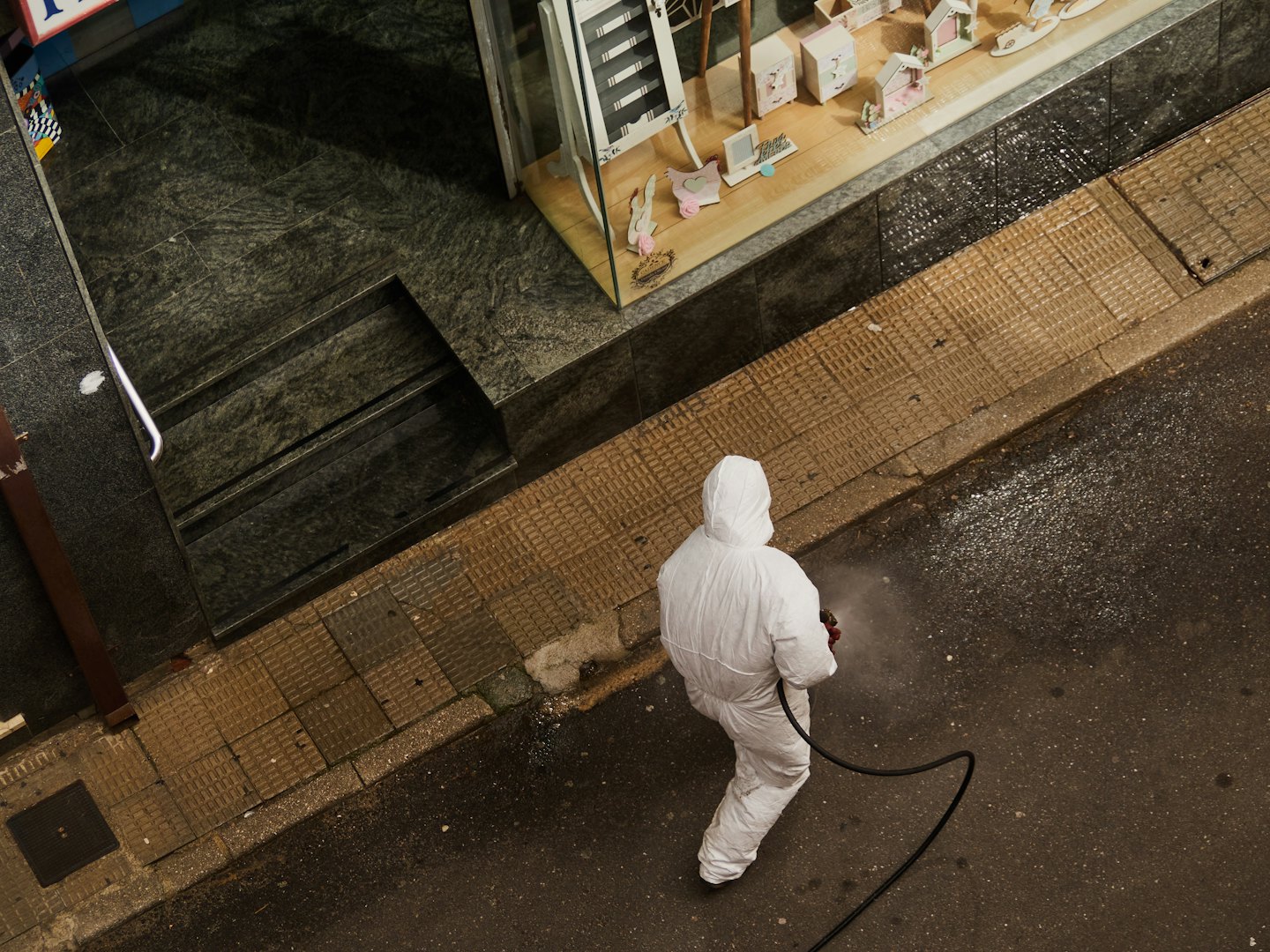 Hazmat suit giymiş bir kişi dükkanın önündeki şehir kaldırımını dezenfekte ediyor.